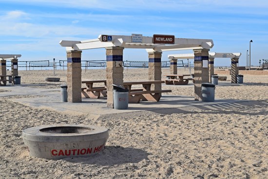 Newland Ramada B sits on a paved path (wheelchair accessible). There are two picnic tables and a fire ring adjacent to the area. Volleyball courts line the sand several yards behind the picnic area.